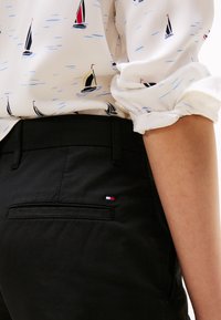 I pantaloni neri presentano una tasca sul retro con un piccolo logo ricamato. La blusa ha uno sfondo bianco con motivi di barche a vela blu e neri.