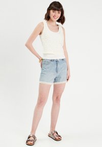 Débardeur en crochet blanc avec un motif floral, associé à un short en denim bleu clair avec une bordure en dentelle. Des sandales métalliques marron complètent le look.
