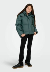 Grön vadderad jacka med dragkedja framtill, stor krage och tryckknappar. Matchas med svarta figursydda byxor och svarta sneakers med gummisula.