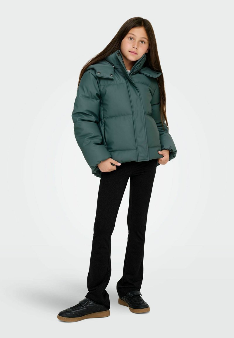 Grön vadderad jacka med dragkedja framtill, stor krage och tryckknappar. Matchas med svarta figursydda byxor och svarta sneakers med gummisula.