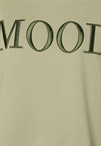 T-shirt en coton vert avec le mot "MOOD" brodé en vert foncé. Le tissu a une texture lisse et une coupe décontractée.