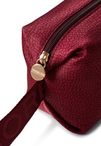 Small burgundy bag with a textured fabric, gold zipper, and a round logo tag. Features a sturdy strap with embossed lettering.