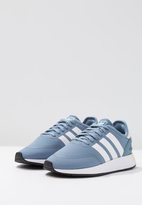 Ljusblå atletiska sneakers med tre vita ränder, texturerat tyg, vit mellansula och svart gummisul. Snörningsdesign.