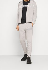 Light gray tracksuit with a zip-up jacket and matching joggers, featuring black logo text. Black t-shirt underneath and black sneakers.