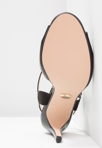 Chaussure noire à talon haut inclinée vers le haut, montrant une semelle beige lisse avec un petit emblème doré de la marque près de la pointe du talon.
