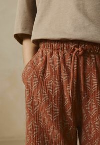 Pantalon couleur rouille avec une ceinture élastique, arborant un motif de feuilles géométriques en blanc et beige. Tissu doux et texturé avec des poches.