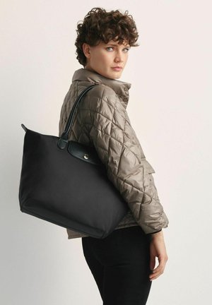 Duża czarna torba tote o błyszczącej fakturze i czarnych skórzanych uchwytach, połączona z pikowaną beżową kurtką, na neutralnym tle.