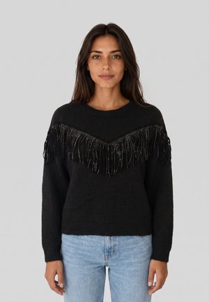 Donna con capelli lunghi e scuri che indossa un maglione nero con frange di perline sul petto e jeans azzurri, in piedi davanti a uno sfondo semplice.