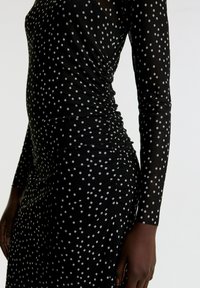 Schwarzes Kleid mit langen Ärmeln, das ein gerafftes Design und weiße Polka-Dots aufweist. Der Stoff erscheint leicht und leicht transparent.