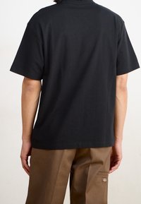 Svart bomullströja med kort ärm och klassisk passform, med en enfärgad baksida och kontrasterande bruna byxor med sidficka.