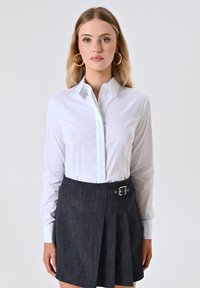 Camicia bianca a maniche lunghe con colletto a punta, abbinata a una gonna plissettata in denim scuro con un dettaglio in fibbia argentata.