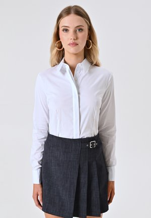 Camicia bianca a maniche lunghe con colletto a punta, abbinata a una gonna plissettata in denim scuro con un dettaglio in fibbia argentata.