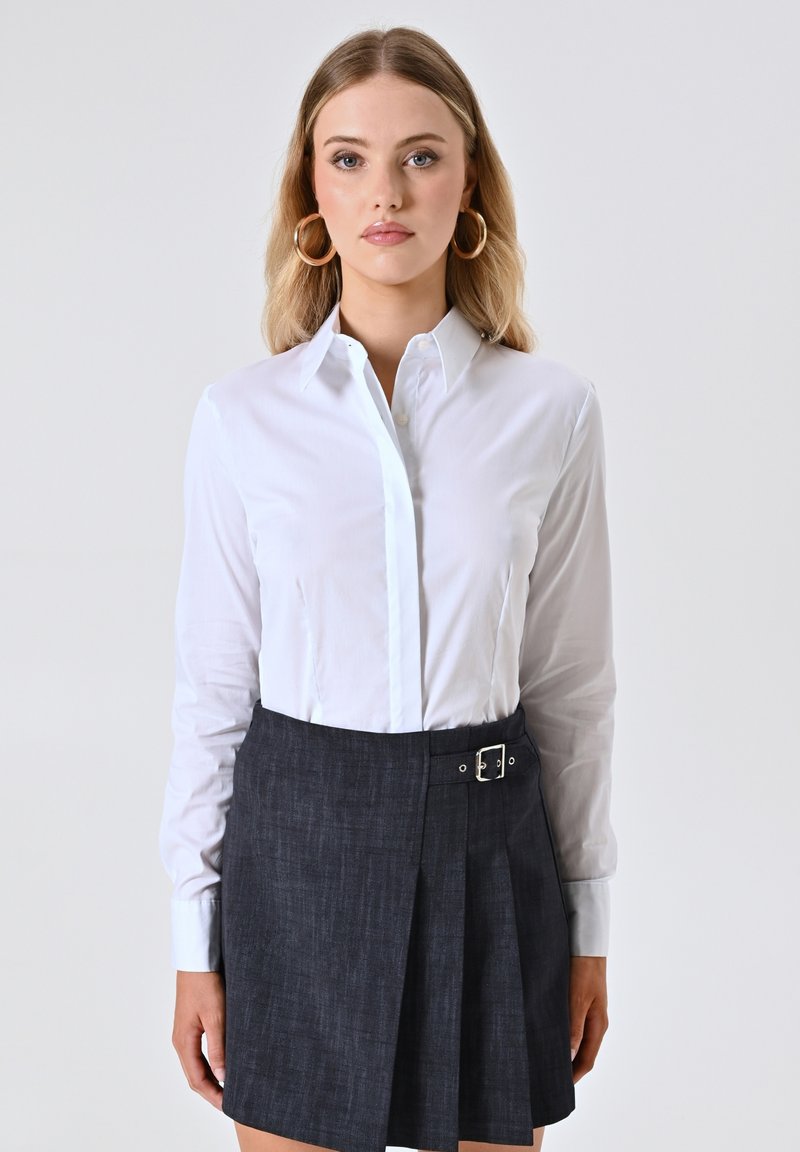 Camicia bianca a maniche lunghe con colletto a punta, abbinata a una gonna plissettata in denim scuro con un dettaglio in fibbia argentata.