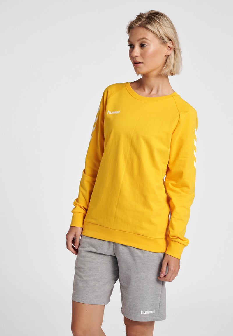 Gul sweatshirt med lång ärm, rundad krage och vit logotyp; matchad med grå shorts. Slätt tyg, atletisk passform och kontrasterande detaljer.