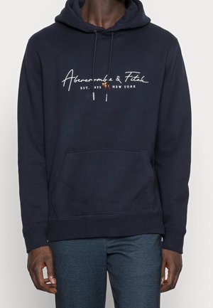 Mężczyzna ubrany w granatową bluzę z kapturem Abercrombie & Fitch z przednią kieszenią i ściągaczem przy kapturze, w połączeniu z ciemnymi, teksturowanymi spodniami.