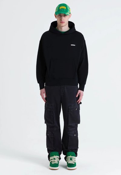 Sweat-shirt noir à capuche avec poche avant, associé à un pantalon cargo noir doté de plusieurs poches. Baskets vertes avec de grosses lacets blancs.