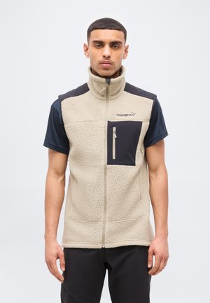 Junger Mann, der beige Fleece-Weste mit Reißverschluss und schwarzer Brusttasche sowie schwarzem Kurzarmhemd trägt, vor hellem, einfarbigem Hintergrund stehend.