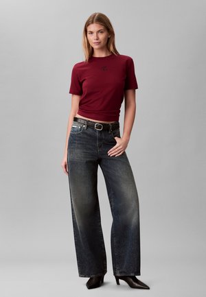 Jeune femme portant un t-shirt bordeaux ajusté à manches courtes, un jean noir délavé à jambes larges, une ceinture noire et des bottines noires à bouts pointus.