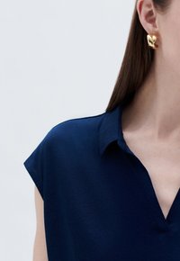 Top blu navy a maniche corte con collare a V. Orecchini geometrici dorati con finitura lucida. I capelli sono lunghi e lisci.