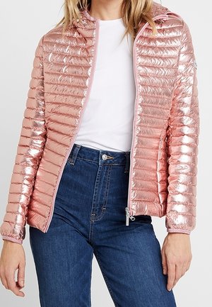 Vrouw draagt een glanzend roze doorgestikte jas over een wit hemd en hooggetailleerde blauwe jeans, staat met handen ontspannen langs haar zij.