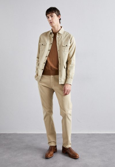 Light beige jacket with two chest pockets, paired with a brown sweater and matching beige pants. Brown leather boots complete the look.
