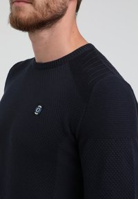 Pull en maille bleu marine avec motifs texturés, col côtelé et détails aux épaules. Présente un petit logo sur la poitrine.