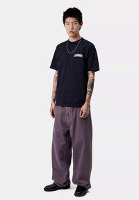Czarna bawełniana koszulka z białym logo "Carhartt", w połączeniu z luźnymi, jasnopurpurowymi spodniami i czarnymi butami. Model ma tatuaże i krótkie, sterczące włosy.