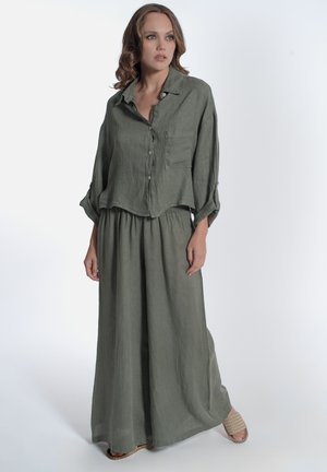 Donna che indossa una camicia larga di lino verde oliva a maniche lunghe con bottoni e pantaloni a gamba larga con sandali beige intrecciati, in piedi su sfondo bianco.