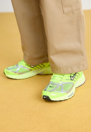 Sneakers atletici verde neon con mesh e accenti argento, dotati di un logo nero, abbinati a pantaloni beige chiaro, su una superficie gialla.