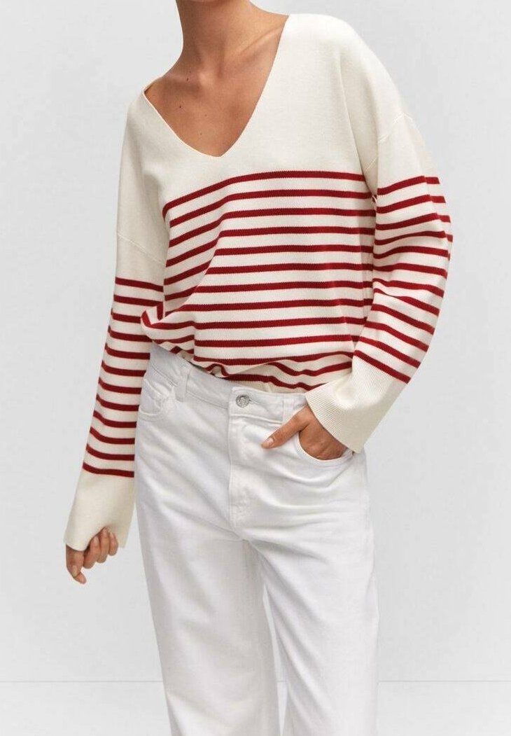 Personne portant un pull blanc à col en V avec des rayures horizontales rouges et un pantalon blanc, une main dans la poche, devant un fond uni.