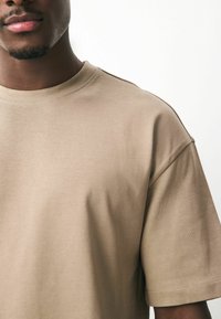 T-shirt beige à manches courtes en coton doux avec un col ras du cou, présentant une coupe décontractée et un détail de couture minimal sur l'épaule.