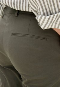 Dunkelolivgrüne Hosen mit glatter Textur, ausgestattet mit einer Gesäßtasche mit dezentem genähtem Akzent. Kombiniert mit einem gestreiften Hemd.