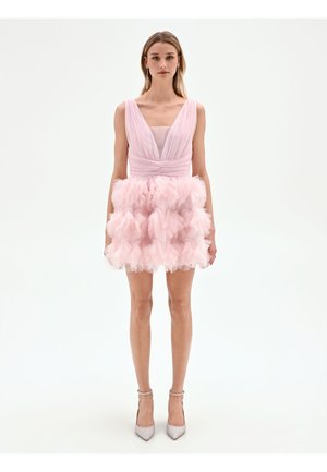Jeune femme portant une robe sans manches rose pâle avec un décolleté en V plongeant et une jupe en tulle à volants superposés, associée à des talons légers à bride de cheville.