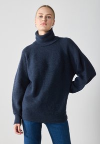 Pullover oversized de culoare bleumarin închis, dintr-un material tricotat texturat, cu mâneci lungi și o croială relaxată. Purtați-l cu blugi albaștri.