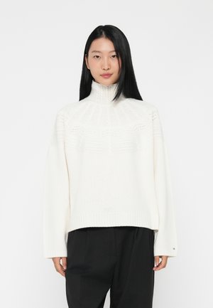 Pull en tricot blanc à col montant avec un design texturé au niveau de l'emmanchure, ourlet et poignets côtelés, associé à un pantalon noir.