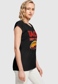 Sort t-shirt med rund halsudskæring, kortærmede, der har fed rød tekst "TACO DIRTY TO ME" og et farverigt taco-grafik.