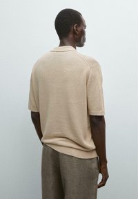 Beige knitted short-sleeve sweater with a ribbed collar and hem, paired with light brown textured trousers, viewed from the back.