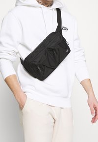 Man i vit Levi's-hoodie och krämfärgade byxor med svart axelväska med flera dragkedjefickor.