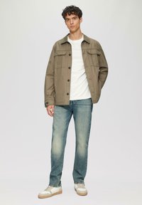 Olijfgroen, knoopjack met twee voorzakken, gecombineerd met lichtblauwe jeans en witte sneakers met tan zolen. Casual pasvorm en modern ontwerp.