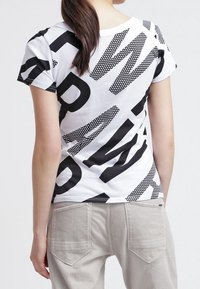 Bílý tričko s velkým černým textem a tečkovanými vzory v hravém uspořádání. Krátké rukávy a kulatý výstřih.