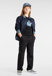 Cazadora bomber azul marina con detalles en azul claro, camiseta gráfica negra, pantalones negros y zapatillas de cuadro blanco y negro. Gorra azul.