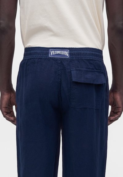 Personne à la peau foncée portant une chemise blanche et un pantalon bleu marine avec une taille élastique et une poche arrière portant le label Vilebrequin.