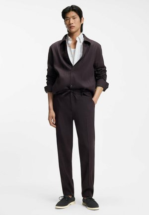 Giovane uomo in piedi con una mano in tasca, indossa pantaloni scuri a taglio comodo, una camicia scura abbinata sopra una maglietta bianca e scarpe nere con lacci.