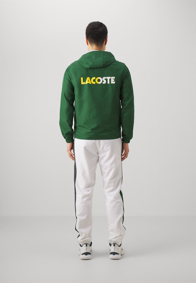 Chaqueta verde con capucha y logo de "LACOSTE" en blanco y amarillo en la parte posterior, combinada con pantalones blancos con franjas laterales negras y zapatillas blancas.
