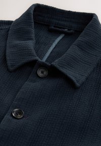 Marineblå skjorte med knapper, fremstillet i et tekstureret stof med vaffelmønster, med krave og en enkelt sort knap foran.