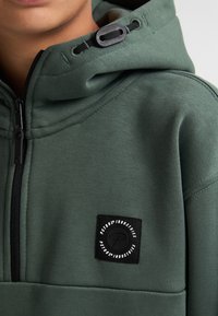 Tmavě zelená mikina s kapucí na šňůrku, zipovým uzávěrem a černým štítkem s bílým textem na levé straně. Hladká textura.