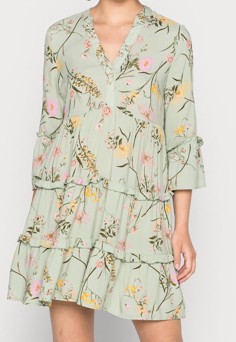 Robe fleurie vert clair avec des manches trois-quarts à volants, une jupe à étages et un décolleté en V, portée par une personne debout avec un bras visible.