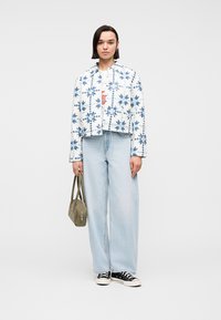 Γυναίκα που φοράει λευκό και μπλε σακάκι με σχέδια, ανοιχτό μπλε τζιν με φαρδιά μπατζάκια, μαύρα αθλητικά παπούτσια, κρατώντας μία χακί τσάντα στο πλάι της.