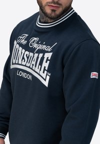 Mann trägt einen navyblauen Lonsdale London Sweatshirt mit weißem gestreiftem Kragen und Logo-Schriftzug auf der Brust und dem Ärmel, zeigt Bart und Nackentattoo.