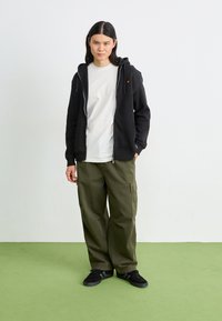 Black zip-up hoodie with a front pocket, paired with olive green cargo pants. White t-shirt underneath, worn with dark sneakers on green floor.
